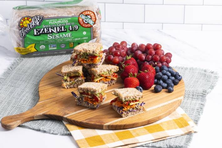Vibrant Veggie Stack Sandwich on Ezekiel 4:9 Sesame Sprouted Whole Grain Bread Colorful veggie sandwich on a cutting board, garnished with berries and grapes, with a package of Food for Life Ezekiel 4:9 bread in the background