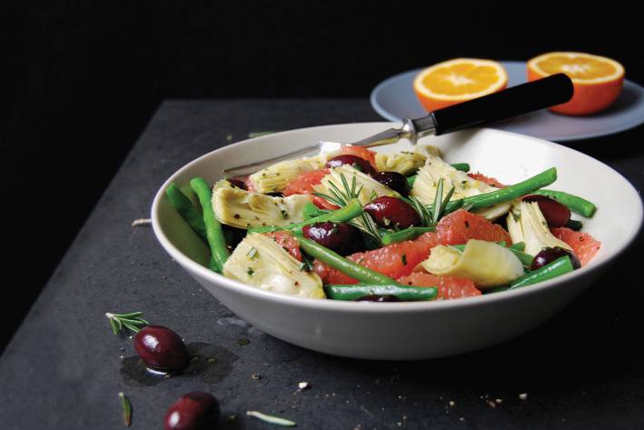 Green Bean, Artichoke, and Grapefruit Salad with Olives & Rosemary Green Bean, Artichoke, & Grapefruit Salad with Olives and Rosemary in a white bowl with a cut orange in the background. Black background.