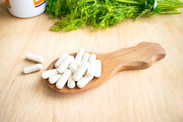Health Benefits of Supplements Vitamins, supplements in spoon on wooden table with green herbs in the background.